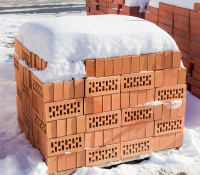 Red perforated bricks with rectangular holes covered snow on a pallet on an outdoor warehouse in winter sunny day
