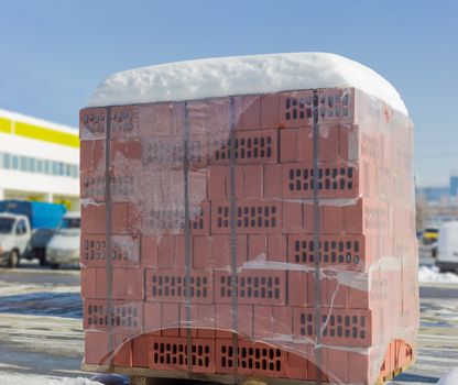 Red perforated bricks packed on a wooden pallet with plastic wrap and covered snow on an outdoor warehouse in winter sunny day
