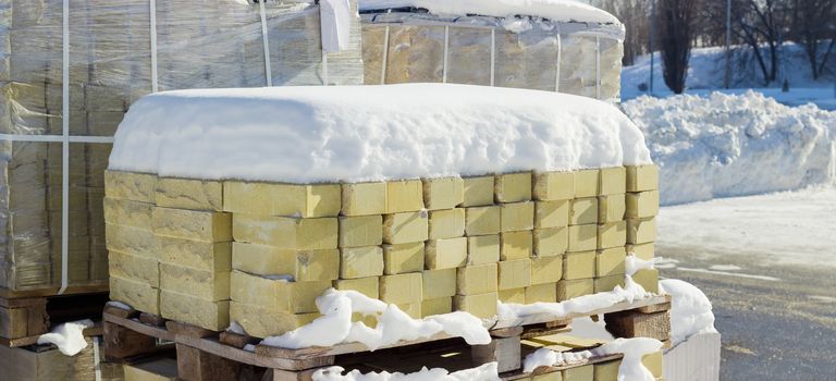 Yellow decorative face bricks covered snow on the wooden pallet against the background of other pallets with bricks on an outdoor warehouse in winter sunny day
