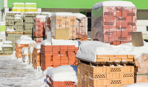 Pallets with different red bricks and concrete masonry units covered snow on an outdoor warehouse in winter sunny day
