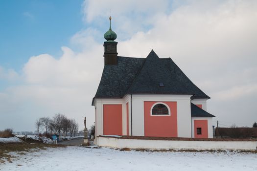 Christian church in village Kninice in Moravia, Czech republic. Snow on the field. Intense cloudy sky. Green tower of the church.