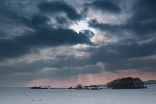 Field covered with snow. Trees and bushes. Mountain in the background. Intense clouds with a winter sun.