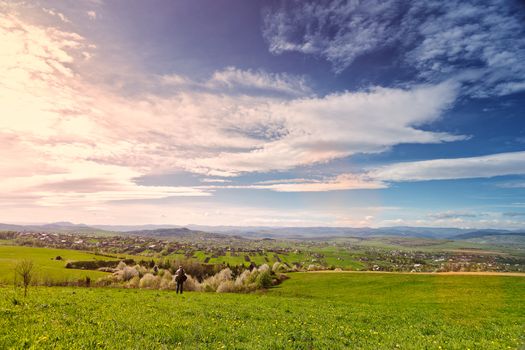 Traveler on green spring meadow. Village on sunny green spring foothills. Orchard blooming on hills. Sunny green blooming spring landscape.