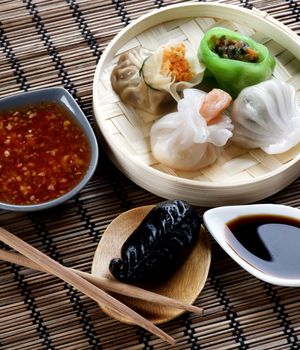 Assorted Dim Sum in Bamboo Steamed Bowl and Igaya  with Seafood on Wooden Plate with Red Chili and Soy Sauces and Chopsticks closeup on Straw Mat background