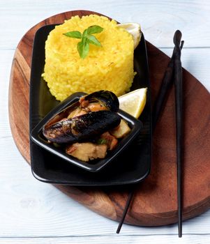Arrangement of Delicious Seafood Curry with Mussels, White Fish and Vegetables and Stack of Saffron Rice with Chopsticks on Serving Board closeup on Wooden background