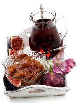 Homemade Homemade Fig Jam in Glass Kremanka with Fresh Fig Fruits, Glass Cup of Tea and Flowers in White Wooden Tray closeup on White background