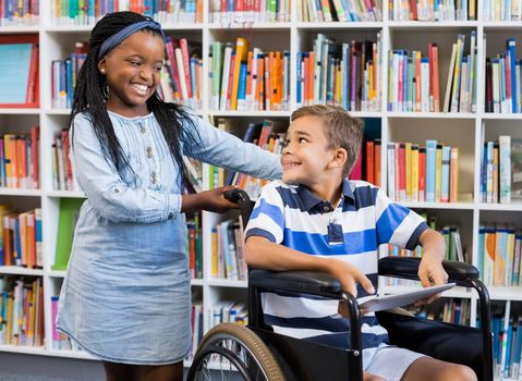Smiling schoolgirl standing with disabled boy on wheelchair in library