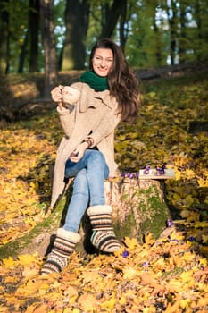 girl with a cup of coffee sitting on a tree stump in autumn Park