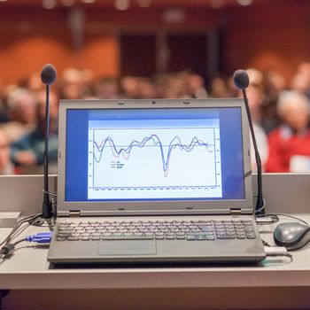Computer and microphone on a podium desk at business event. Blured audience at the conference hall in background. Business and Entrepreneurship concept. Focus on computer.