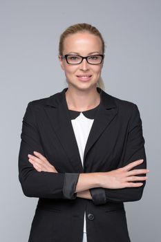 Portrait of beautiful smart young businesswoman in business attire wearin black eyeglasses, standing with arms crossed against gray background.