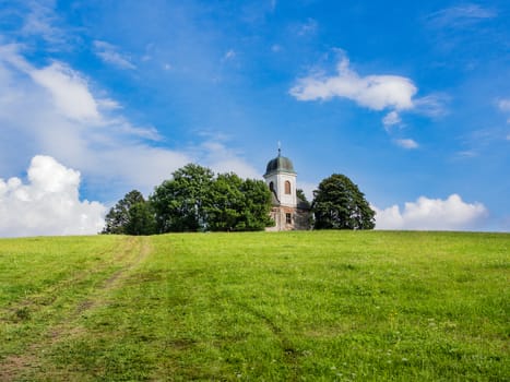 Christian church or chapel with white tower on top of the hill in the forest, horizon between green ground and blue cloudy sky, copy space, relaxing travelling under blue sky