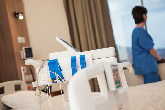 Cardiac stress test equipment in a modern hospital room with a blurred figure of a patient looking through the window.