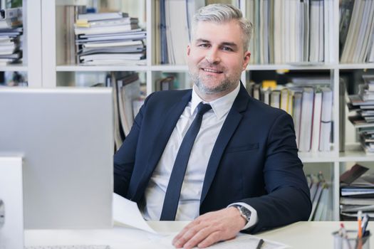 Portrait of mature business man with grey hair in his modern office with computer and many file folders on shelves on background