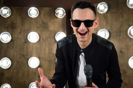 Male singer of rock or pop music dressed in black and sunglasses with microphone performs on scene with lightening projectors on background