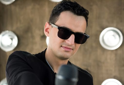 Male singer of rock or pop music dressed in black and sunglasses with microphone performs on scene with lightening projectors on background