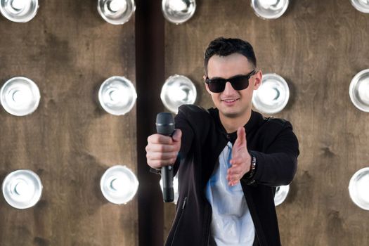 Male singer of rock or pop music dressed in black and sunglasses with microphone performs on scene with lightening projectors on background