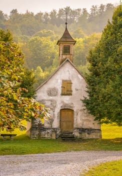 Vertical image with a small medieval church surrounded by trees with autumn colored leaves. Picture taken in a German village.