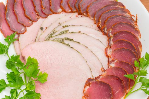 Fragment of white dish with sliced dried pork tenderloin, ham and twigs of parsley closeup
