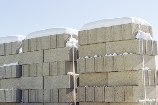 Concrete masonry units packed on pallets and covered snow closeup on an outdoor warehouse in winter sunny day
