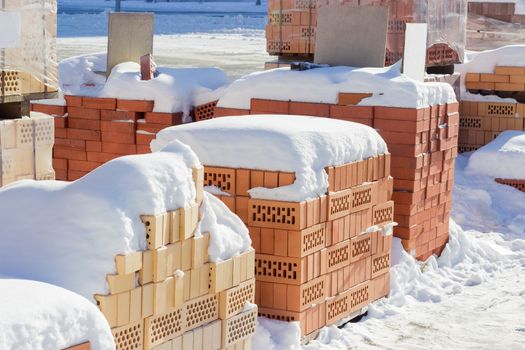 Several pallets of the red perforated bricks with round and rectangular holes covered snow on an outdoor warehouse in winter sunny day
