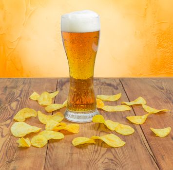 Beer glassware with the lager beer among the potato chips scattered the old darkened by time wooden planks on a background of an yellow wall
