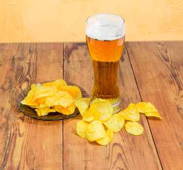 Beer glassware with the lager beer and potato chips on the glass saucer and scattered beside on the old wooden planks on a background of an yellow wall
