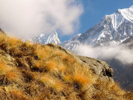 Mountain range with peaks covered with hanging glaciers partly shrouded in clouds with a mountain slope covered with the withered grass in the foreground in the Himalayas
