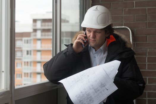 Engineer standing on the background of a new apartment building. House is built. Engineer in a helmet with papers in hand on the phone. The concept of new housing construction, real estate.