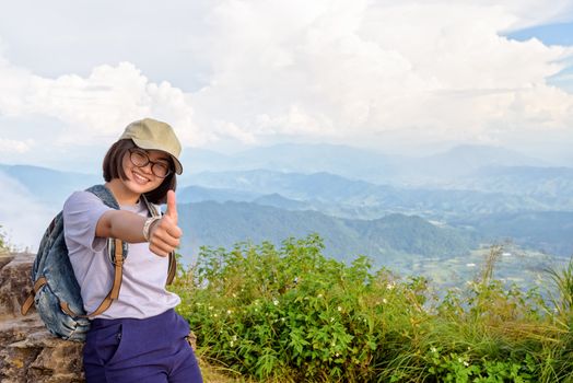 Tourist teens girl hiker wear cap and glasses with backpack poses thumb up smiling happily on high mountain at scenic point of Phu Chi Fa Forest Park in Chiang Rai Province Thailand