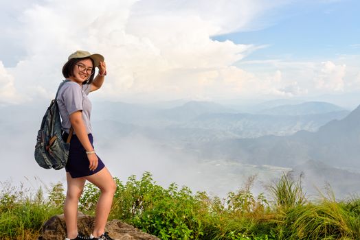 Tourist teens girl hiker with backpack cap and glasses is standing smile and poses happily at high mountain on sky and fog background at scenic point of Phu Chi Fa Forest Park in Chiang Rai, Thailand