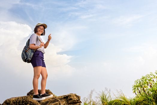 Happy hiker asian cute teens girl with backpack cap and glasses standing smiling poses thumb up on mountain and sky background at Phu Chi Fa Forest Park during sunset in Chiang Rai, Thailand