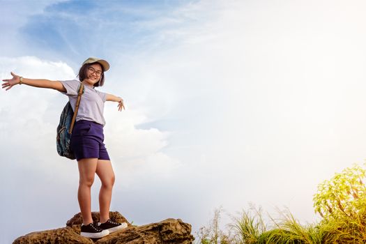 Happy hiker asian cute teens girl with backpack cap and glasses standing smiling poses open arms on mountain and sky background at Phu Chi Fa Forest Park during sunset in Chiang Rai, Thailand