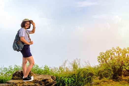 Tourist teens girl hiker with backpack cap and glasses is standing smile and poses happily at high mountain on sky and fog background at scenic point of Phu Chi Fa Forest Park in Chiang Rai, Thailand