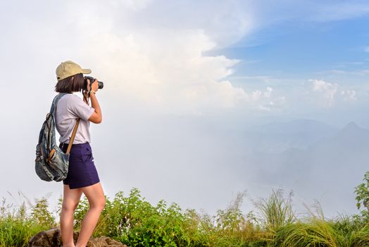 Hiker asian teens girl with digital camera and backpack taking photo beautiful natural of sierra sky clouds and fog during sunset in winter on mountain at Phu Chi Fa Forest Park, Chiang Rai, Thailand
