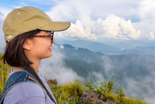 Hiker asian cute teens girl with caps and glasses smiling happily while looking beautiful landscape nature of mountain range and sky during sunset in winter at Phu Chi Fa Forest Park, Chiang Rai, Thailand