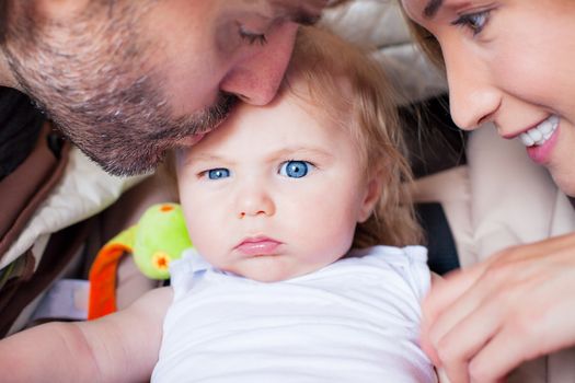 Frowning baby boy with big blue eyes is being kissed and pampered by his parents.