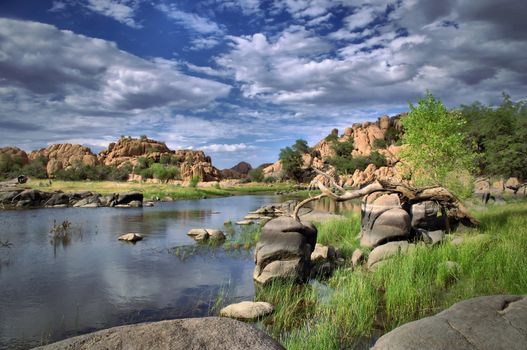 A view from the shore of Watson Lake with blue cloud filled sky and green trees with rock cliffs in Prescott, Arizona.