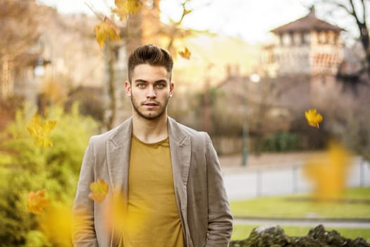 Confident young man standing in the autumn park with ancient castle in background and falling leaves in foreground. Horizontal outdoors shot.