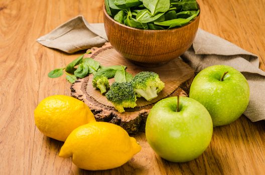 Spring spinach leaves in the bowl, broccoli, lemons and apples on wooden table background