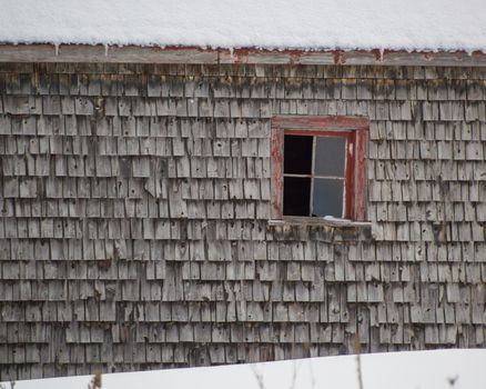 Old weathered cedar shakes shingles barn with red window winter. Snow on the roof and ground.