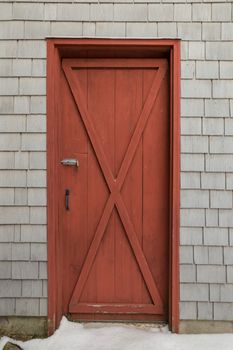 Oxblood red barn style X door on cedar shakes shingles background. Snow on the ground.