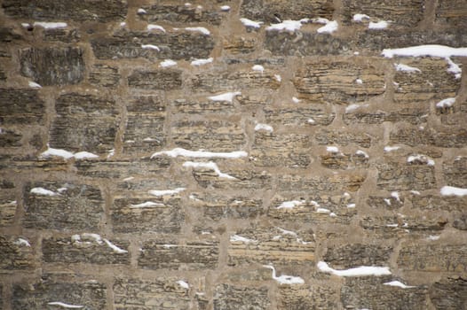 Rugged limestone wall that is uneven and covered with snow in little ledges.  Strata easily seen in the rocks. Rustic and filled with cement mortar.