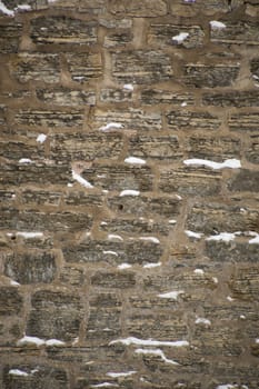 Rugged limestone wall that is uneven and covered with snow in little ledges.  Strata easily seen in the rocks. Rustic and filled with cement mortar.