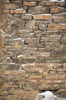 Rugged limestone wall that is uneven and covered with snow in little ledges.  Strata easily seen in the rocks. Rustic and filled with cement mortar.