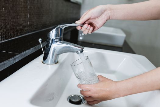 Woman filling a glass of water from a stainless steel or chrome tap or faucet, close up on her hand and the glass with running water and air bubbles.