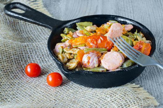 Fried vegetables with chopped sausage in a cast iron pan, a piece on a fork. Gray wood background, burlap and selective focus.