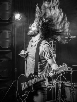 Photo of a young man playing electric guitar on stage and tossing his long hair around.