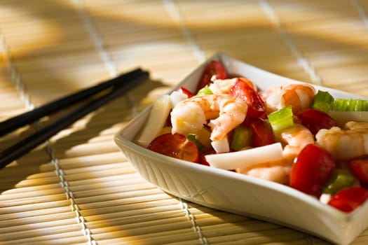 Close up of shrimp salad with squid tomatoes celery inside a white bowl over a bamboo tablecloth and chopsticks