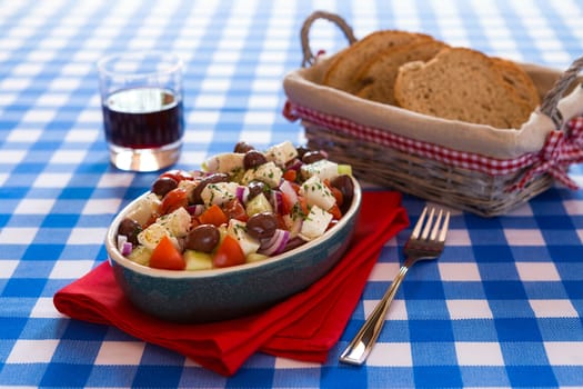 Greek salad with feta cheese olives tomatoes cucumber and onions over a checkered tablecloth with red wine and bread