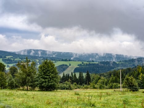Natural cloudy misty landscape. mountain meadow and forests, both deciduous broadleaved and needle coniferous trees, electric overhead power line and horizon covered in clouds and mist, Czech republic, central Europe, Orlicke hory